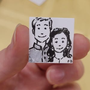 May include: A small, square, hand-drawn illustration of two people, likely a couple, in black ink and watercolor. The artwork is on white paper and held by a hand. The style is simple and charming, with a focus on the faces.