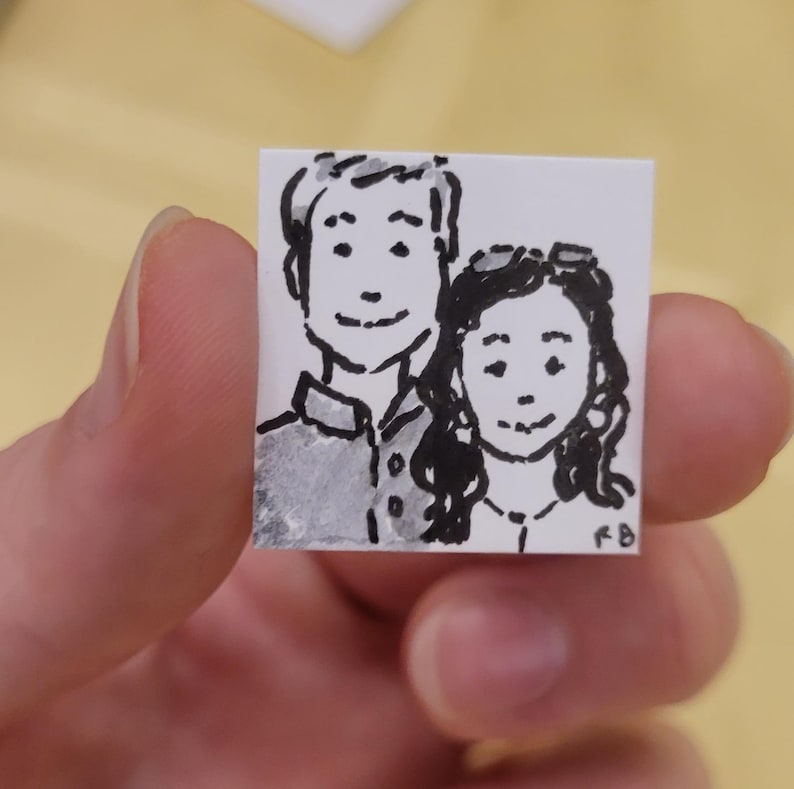 May include: A small, square, hand-drawn illustration of two people, likely a couple, in black ink and watercolor. The artwork is on white paper and held by a hand. The style is simple and charming, with a focus on the faces.
