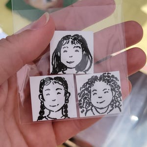 May include: Three small square stickers with black and white illustrations of smiling faces. One has long hair, one has braids, and the third has curly hair. The stickers are in a clear plastic bag.