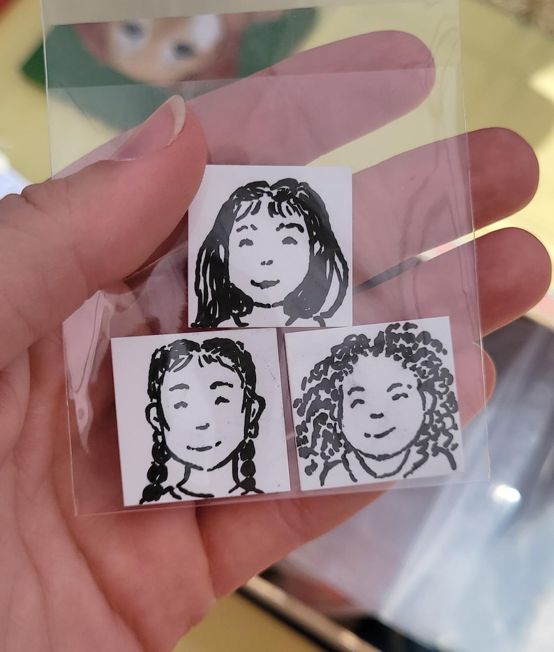 May include: Three small square stickers with black and white illustrations of smiling faces. One has long hair, one has braids, and the third has curly hair. The stickers are in a clear plastic bag.