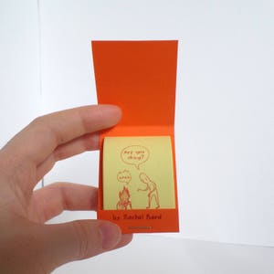 May include: A small, yellow notepad with a red illustration of two stick figures. The text on the notepad reads "Are you okay?" and "Yeah". The notepad is open and has a red cover with the text "by Rachel Bard" on the bottom.
