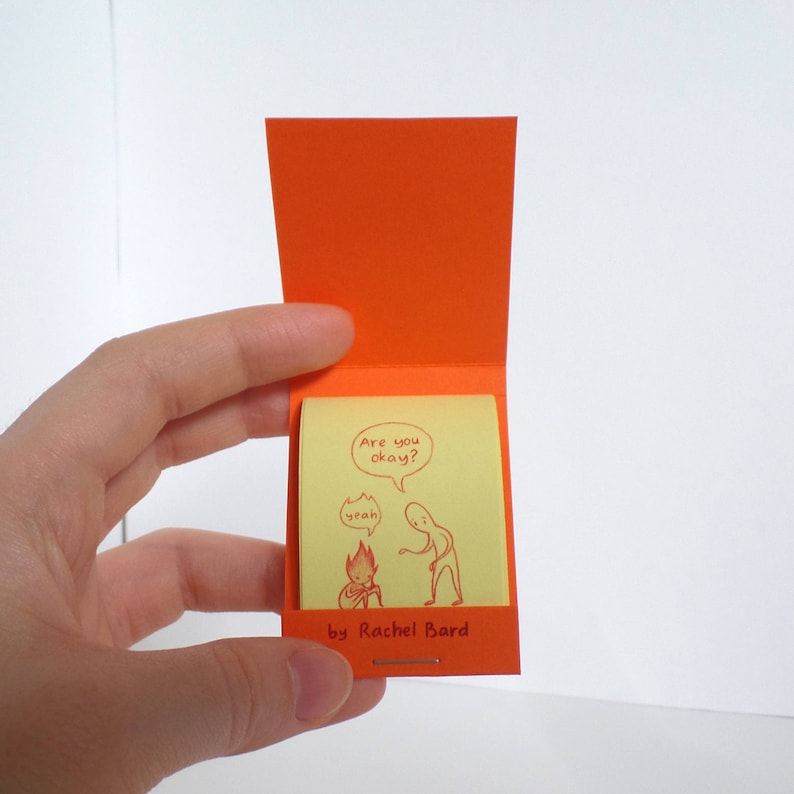 May include: A small, yellow notepad with a red illustration of two stick figures. The text on the notepad reads "Are you okay?" and "Yeah". The notepad is open and has a red cover with the text "by Rachel Bard" on the bottom.