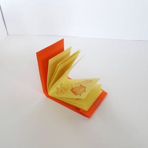 May include: A small, orange-covered book with yellow pages. The pages are folded and the book is open, revealing a few pages with red drawings of leaves and flowers.