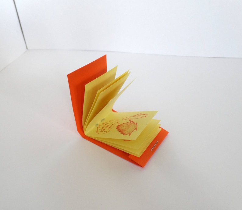 May include: A small, orange-covered book with yellow pages. The pages are folded and the book is open, revealing a few pages with red drawings of leaves and flowers.