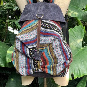 May include: A multi-coloured striped backpack with a grey flap and a brown zipper. The backpack has a woven design and a metal ring on the flap.