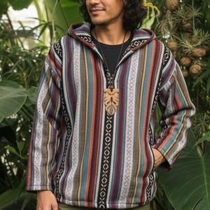 May include: A multi-coloured striped hooded jacket, with shades of red, blue, and brown, worn with olive green trousers and brown boots. A wooden pendant hangs from a black shirt.