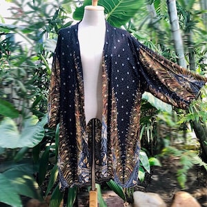 Puede incluir: Un cárdigan negro estilo kimono con un estampado de lunares blancos y un diseño de plumas de pavo real. El cárdigan abierto se exhibe en un maniquí, mostrando sus mangas fluidas y detalles intrincados. La prenda está hecha de una tela ligera.