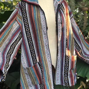 K&ouml;nnte beinhalten: Eine bunt gestreifte Jacke mit Kapuze und zwei Vordertaschen. Die Jacke hat vertikale Streifen in verschiedenen Farben, darunter Rot, Blau, Gelb und Schwarz. Die Jacke wird auf einer Schaufensterpuppe pr&auml;sentiert.
