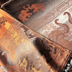 May include: A brown and gold silk scarf with an intricate elephant design. The scarf has a fringe border and is made of a soft, luxurious fabric.
