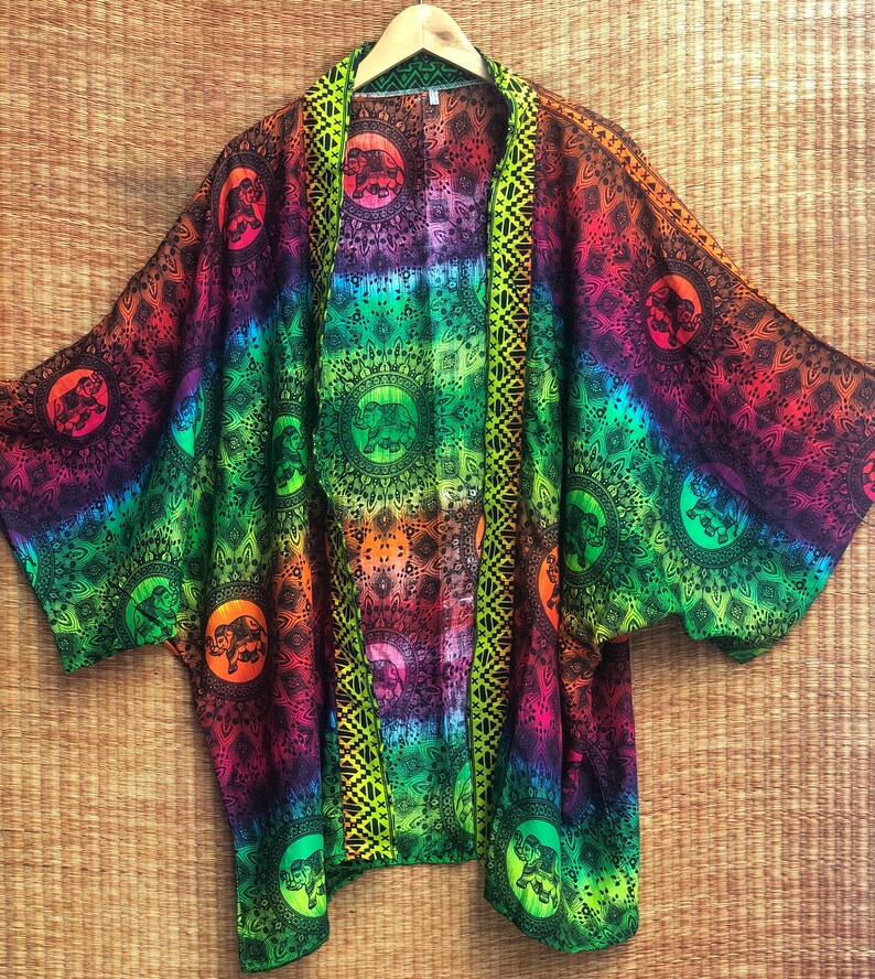 Peut inclure: Un kimono tie-dye arc-en-ciel avec un motif mandala noir et vert repr&eacute;sentant des &eacute;l&eacute;phants. Le kimono est orn&eacute; d'une bordure verte et d'une bordure noire et jaune sur le devant.