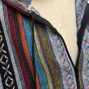 K&ouml;nnte beinhalten: Nahaufnahme einer farbenfrohen, gestreiften Jacke mit Kapuze. Die Jacke hat einen schwarzen Rei&szlig;verschluss und einen Kordelzug mit einer Holzperle. Der Stoff hat eine gewebte Textur mit vertikalen Streifen in verschiedenen Farben.