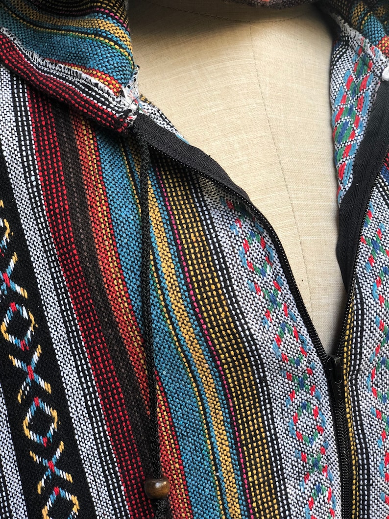 K&ouml;nnte beinhalten: Nahaufnahme einer farbenfrohen, gestreiften Jacke mit Kapuze. Die Jacke hat einen schwarzen Rei&szlig;verschluss und einen Kordelzug mit einer Holzperle. Der Stoff hat eine gewebte Textur mit vertikalen Streifen in verschiedenen Farben.