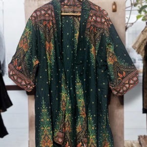Puede incluir: Un kimono abierto de color verde oscuro con un patrón repetitivo de motivos botánicos naranjas y amarillos, bordes geométricos intrincados y pequeños puntos blancos en toda la tela, colgado en una percha de madera.