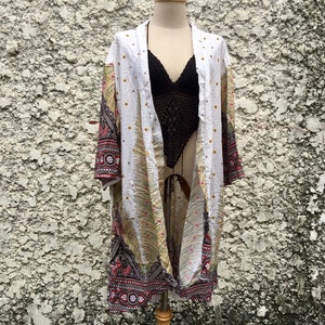 May include: White kimono with a colourful paisley pattern and gold accents. The kimono is open at the front and has long sleeves. It is displayed on a wooden mannequin.