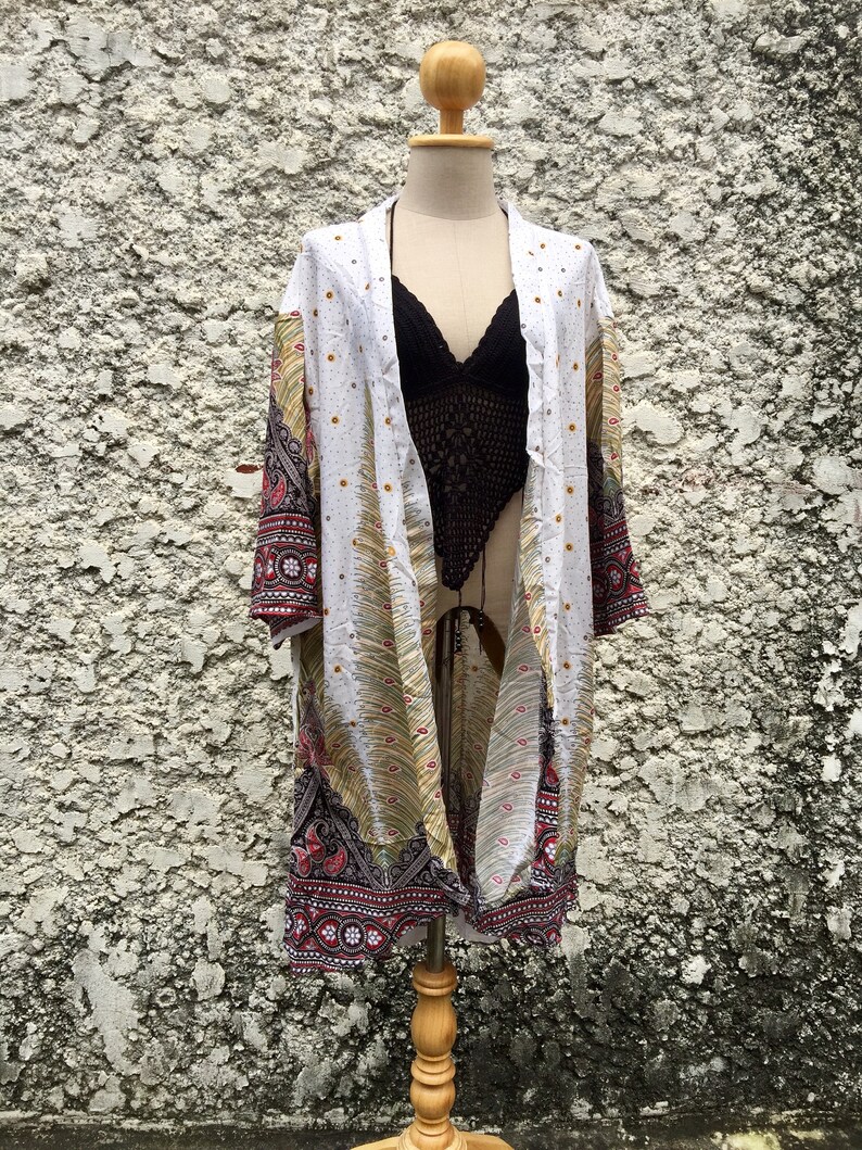 May include: White kimono with a colourful paisley pattern and gold accents. The kimono is open at the front and has long sleeves. It is displayed on a wooden mannequin.