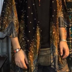 Peut inclure: Kimono noir à pois blancs avec un motif de plumes dorées et marron. Il est porté sur un haut noir et un short à motifs. Un collier de perles et un bracelet complètent l'ensemble. Style bohème.