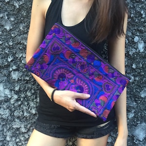May include: A purple and blue embroidered clutch with floral and geometric patterns. The clutch has a black zipper and is held by a person.