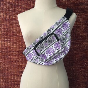 May include: A white fanny pack with a purple and black elephant print. The fanny pack has a zippered pocket on the front and a black adjustable strap.