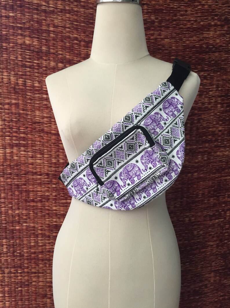 May include: A white fanny pack with a purple and black elephant print. The fanny pack has a zippered pocket on the front and a black adjustable strap.
