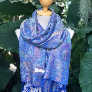 Peut inclure: Une écharpe bleue à motifs paisley avec une bordure à franges. L'écharpe est drapé autour d'un mannequin en bois. L'étiquette sur l'écharpe indique "PASCHAKALIKA".