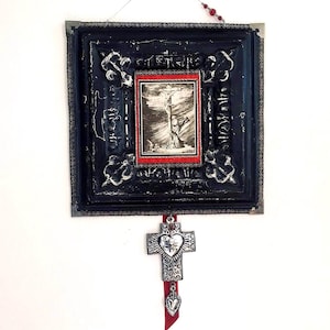 May include: A black framed picture with a red border and a vintage image of a person on a cross. A silver cross with a heart charm hangs from the bottom of the frame with a red ribbon.