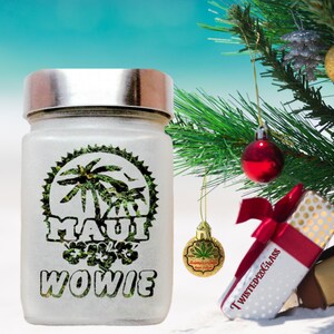 Puede incluir: Un tarro de vidrio blanco con una tapa plateada. El tarro tiene un dise&ntilde;o verde y blanco con las palabras "Maui Wowie" impresas en &eacute;l. El tarro est&aacute; sentado en una playa de arena blanca con un &aacute;rbol de Navidad en el fondo.