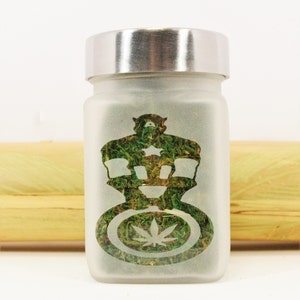 May include: A frosted glass jar with a silver lid, filled with green plant matter. The jar features a crown design with a cannabis leaf emblem. The background is white with a wooden element.