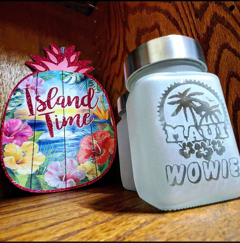 Puede incluir: Un tarro de vidrio blanco esmerilado con tapa plateada. El tarro tiene un dise&ntilde;o en blanco y negro que dice "Maui Wowie" con una palmera y flores. El tarro est&aacute; sentado sobre una superficie de madera junto a un letrero en forma de pi&ntilde;a rosa y verde que dice "Island Time".