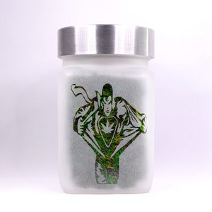May include: A frosted glass jar with a silver lid. The jar has a green and black silhouette of a superhero with a marijuana leaf on their chest.