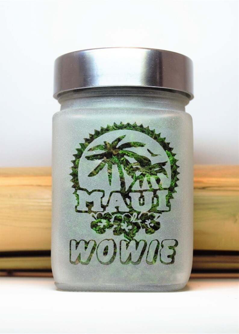 Puede incluir: Un tarro de vidrio esmerilado con tapa plateada. El tarro tiene un dise&ntilde;o verde con palmeras y las palabras "Maui Wowie" en &eacute;l.