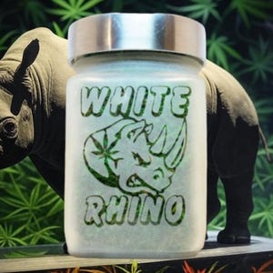 May include: A glass jar with a silver lid, labeled "WHITE RHINO" in green text with a rhino head logo. The jar is in front of a rhino statue and cannabis plants.