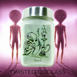 May include: A frosted glass jar with a silver lid features a green alien smoking a hookah. The jar has the text "TWISTED420GLASS" printed on the bottom. Two gray alien figures stand on either side of the jar.