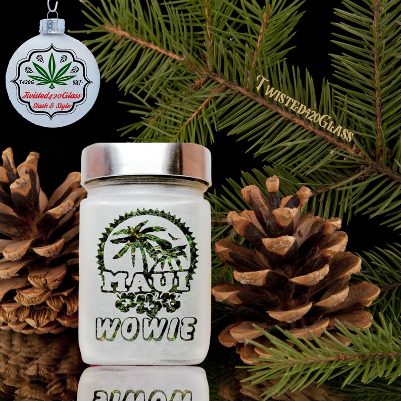 Puede incluir: Tarro de vidrio blanco con un dise&ntilde;o de hoja de marihuana verde y las palabras "Maui Wowie" impresas en &eacute;l. El tarro tiene una tapa plateada y est&aacute; sentado en una mesa con pi&ntilde;as y una rama de &aacute;rbol de Navidad.