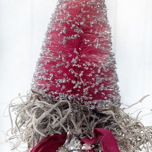 May include: A pink bottle brush Christmas tree with silver glitter, nestled in a bed of dried moss, adorned with a silver metal ornament and a pink ribbon.