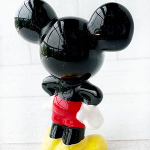 May include: A glossy black Mickey Mouse figurine with large, rounded ears, red shorts, white gloves, and yellow shoes. The figurine is standing upright, with a cheerful expression, and is a collectible item.