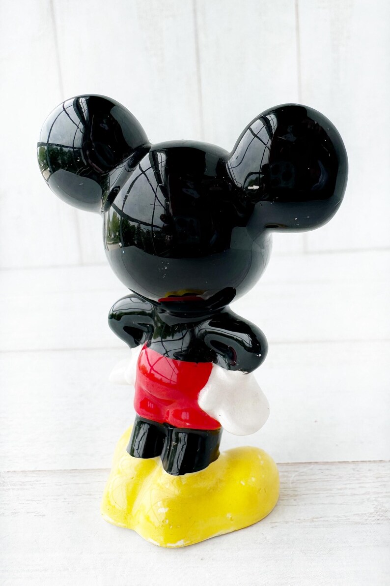 May include: A glossy black Mickey Mouse figurine with large, rounded ears, red shorts, white gloves, and yellow shoes. The figurine is standing upright, with a cheerful expression, and is a collectible item.