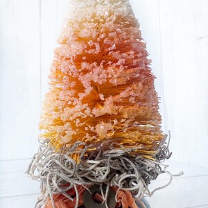May include: A small, orange, white, and yellow bottle brush tree with a brown base and a rusty key. The tree is decorated with a brown, white, and orange ribbon.