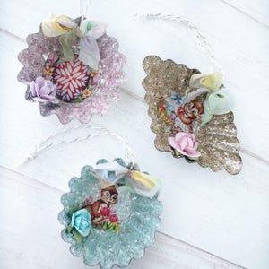 May include: Three vintage-style ornaments with scalloped edges. Each ornament features a different design: a floral pattern, a cartoon chipmunk, and a cartoon chipmunk with a basket. They are adorned with ribbons and small fabric flowers, and have a string for hanging.