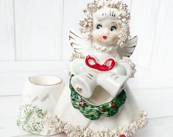 May include: White ceramic angel figurine holding two bells with a red bow. The angel is wearing a white dress with a green wreath around her waist. The figurine is a candle holder with a separate base.