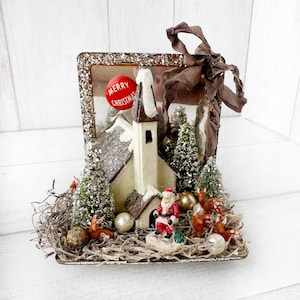 May include: A Christmas-themed decorative piece featuring a church, Santa Claus, deer, and snow-covered trees. The church has a white exterior and brown roof. A red ornament with "Merry Christmas" is visible. The piece is accented with a brown ribbon and sits on a rectangular base.