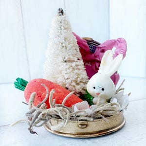 May include: A vintage Easter-themed decorative piece featuring a white bunny, a red carrot, and a white bottle brush tree. The bunny has a smiling face and is nestled in a gold-colored container with faux straw. A pink bow adds a touch of color.
