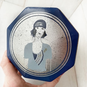 May include: A blue and silver octagonal tin box with a vintage illustration of a woman wearing a cloche hat and a beaded necklace.