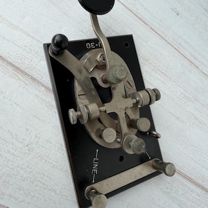 May include: Vintage telegraph key with a black base and silver-toned metal components. The key features a black knob, a lever, and several adjustment screws. The text "LINE" and "J-38" are visible on the base.