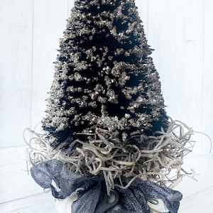 May include: A small black bottle brush Christmas tree decorated with silver glitter, nestled in a bed of dried moss and a black ribbon with a vintage key.