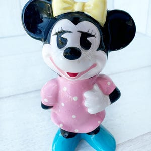 May include: A ceramic figurine of a cartoon character with black ears, a yellow bow, and a pink dress with white polka dots. The character has black eyes, a red mouth, and blue shoes. The figurine is standing on a white surface.