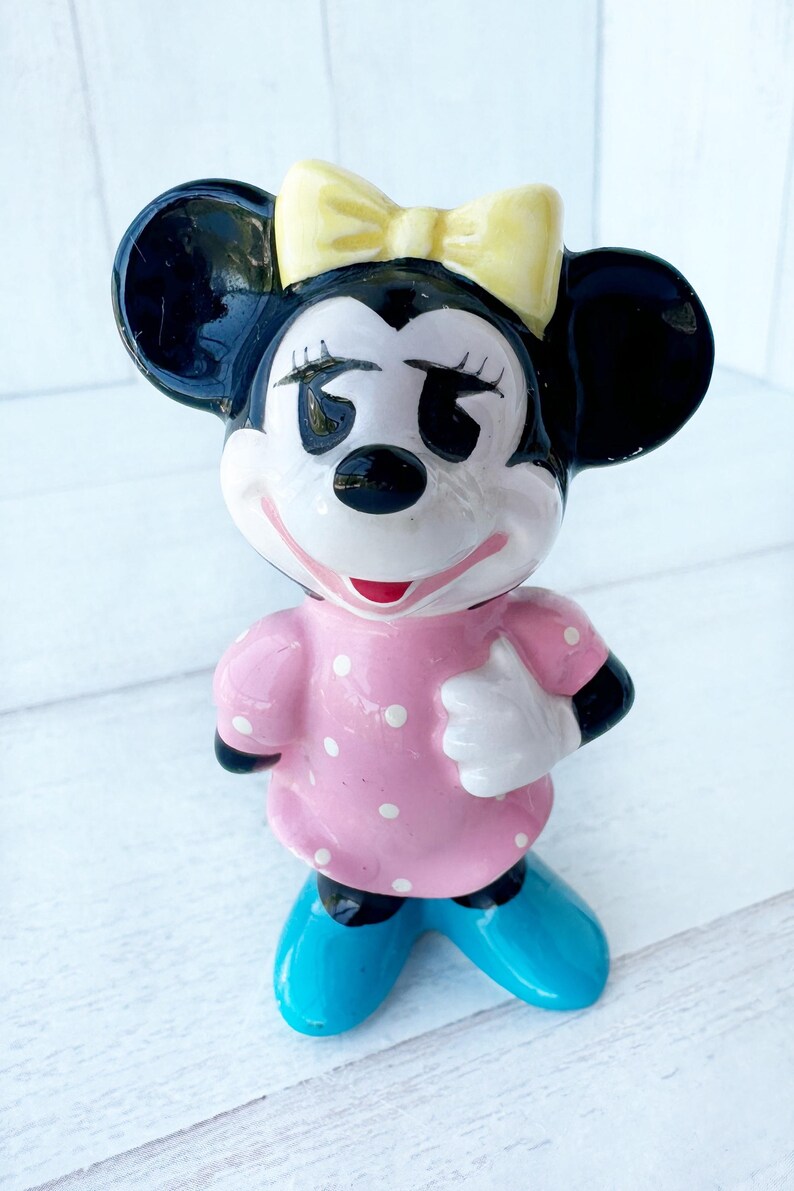 May include: A ceramic figurine of a cartoon character with black ears, a yellow bow, and a pink dress with white polka dots. The character has black eyes, a red mouth, and blue shoes. The figurine is standing on a white surface.