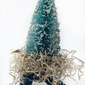 May include: A small teal blue bottle brush Christmas tree with silver glitter, nestled in a bed of beige dried moss, accented with a teal blue ribbon and iridescent jewels.