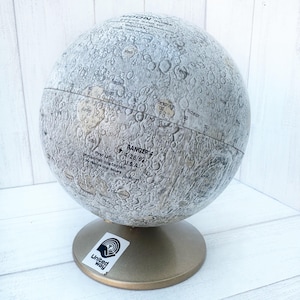 May include: A vintage moon globe with a detailed surface texture, showing craters and other lunar features. The globe is mounted on a gold-colored stand and features the text "RANGER 4 4/26/62 U.S.A." and "First U.S. unmanned spacecraft to reach the Moon." The globe also has a United Way sticker on the stand.
