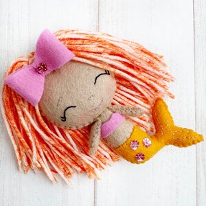 May include: A felt mermaid doll with orange and white yarn hair, a pink bow, and a yellow tail with sequins.