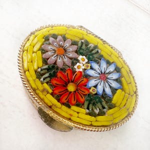May include: A small oval-shaped trinket box with a floral mosaic design. The box is made of metal with a gold finish and features a hinged lid. The mosaic design includes yellow, orange, red, blue, white, and green glass tiles.